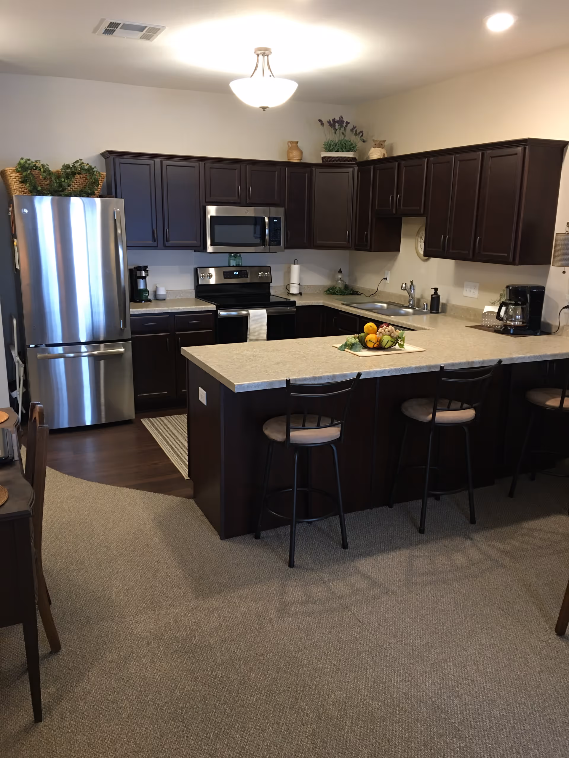 Modern kitchen with dark wood cabinets, stainless steel refrigerator, stove, and microwave. A kitchen island with a beige countertop has three bar stools and a fruit centerpiece. The floor is carpeted with a small area of wood flooring near the appliances. The kitchen is well-lit with a ceiling light fixture.