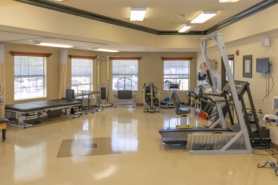 A bright rehabilitation/exercise room with therapy tables, exercise machines, and large windows.