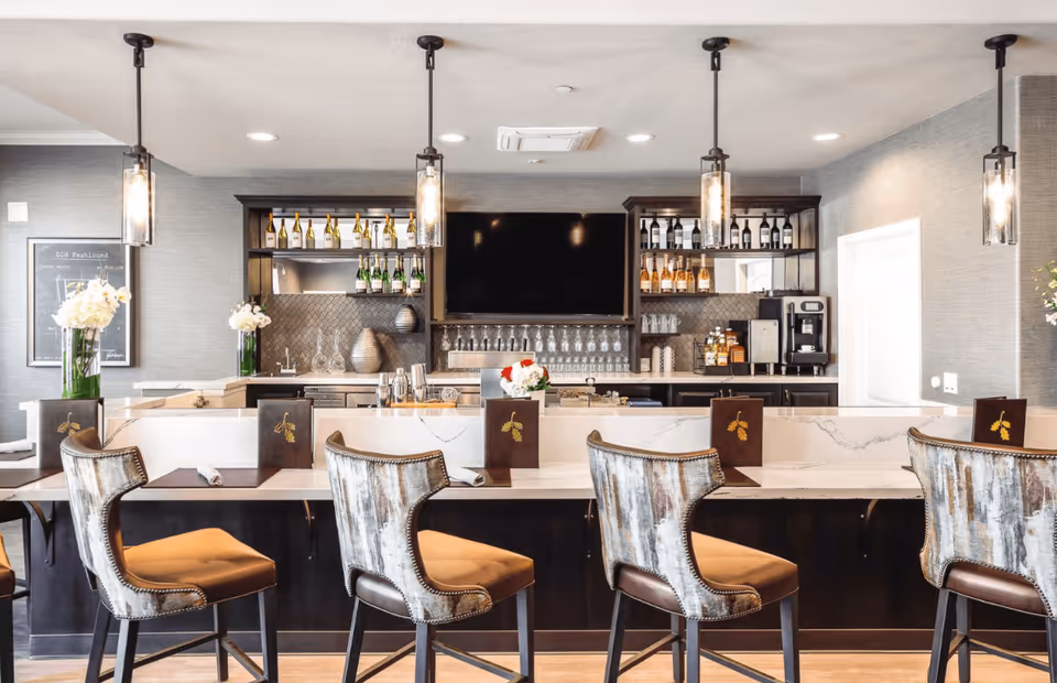 A modern bar area with four high-back chairs lined up along a marble countertop. Behind the counter, there are shelves stocked with wine bottles, glasses, and a coffee machine. Three pendant lights hang from the ceiling, illuminating the space. The decor includes vases with white flowers and a large flat-screen TV mounted on the wall.