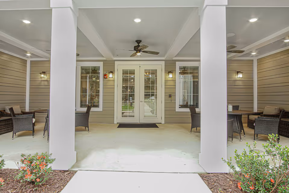 Covered outdoor seating area with wicker chairs and tables on either side of a double glass door entrance. The area has beige siding walls, white columns, ceiling fans, and wall-mounted lights. There are small bushes with red flowers in the foreground.