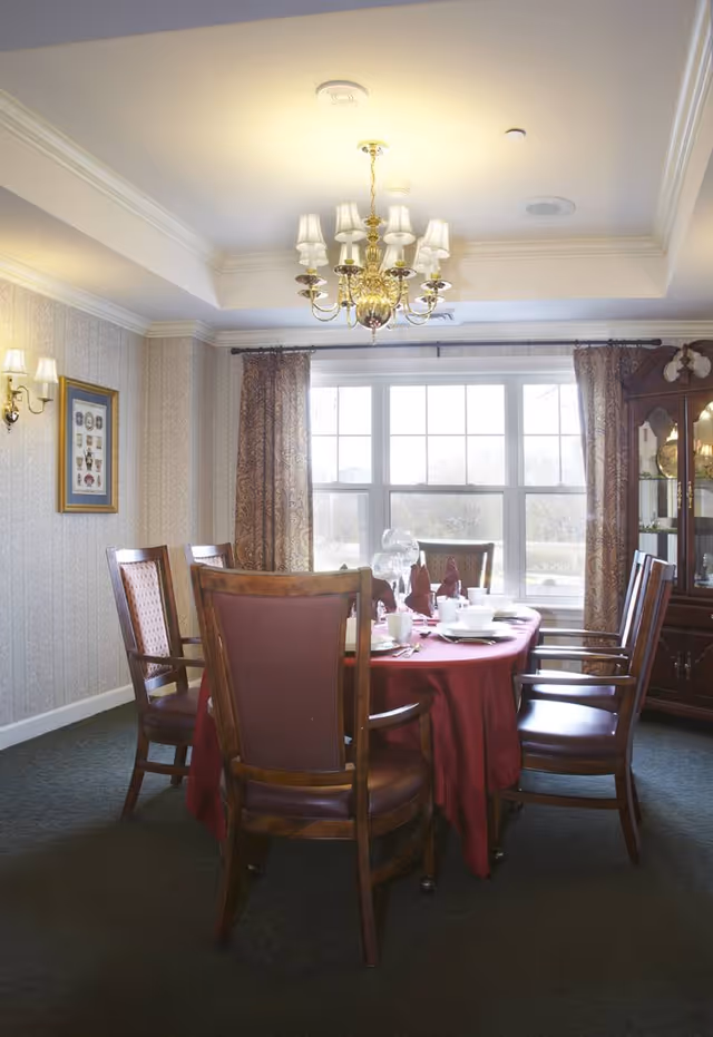 A dining room with a round table covered by a red tablecloth, set with white dishes, glasses, and folded napkins. Six wooden chairs with cushioned seats surround the table. A large window with patterned curtains allows natural light into the room. A chandelier hangs from the ceiling, and a wooden china cabinet is visible in the corner.