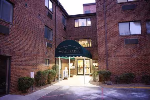 Brick building entrance with a green awning reading Sunridge at Palisades, surrounded by shrubs.