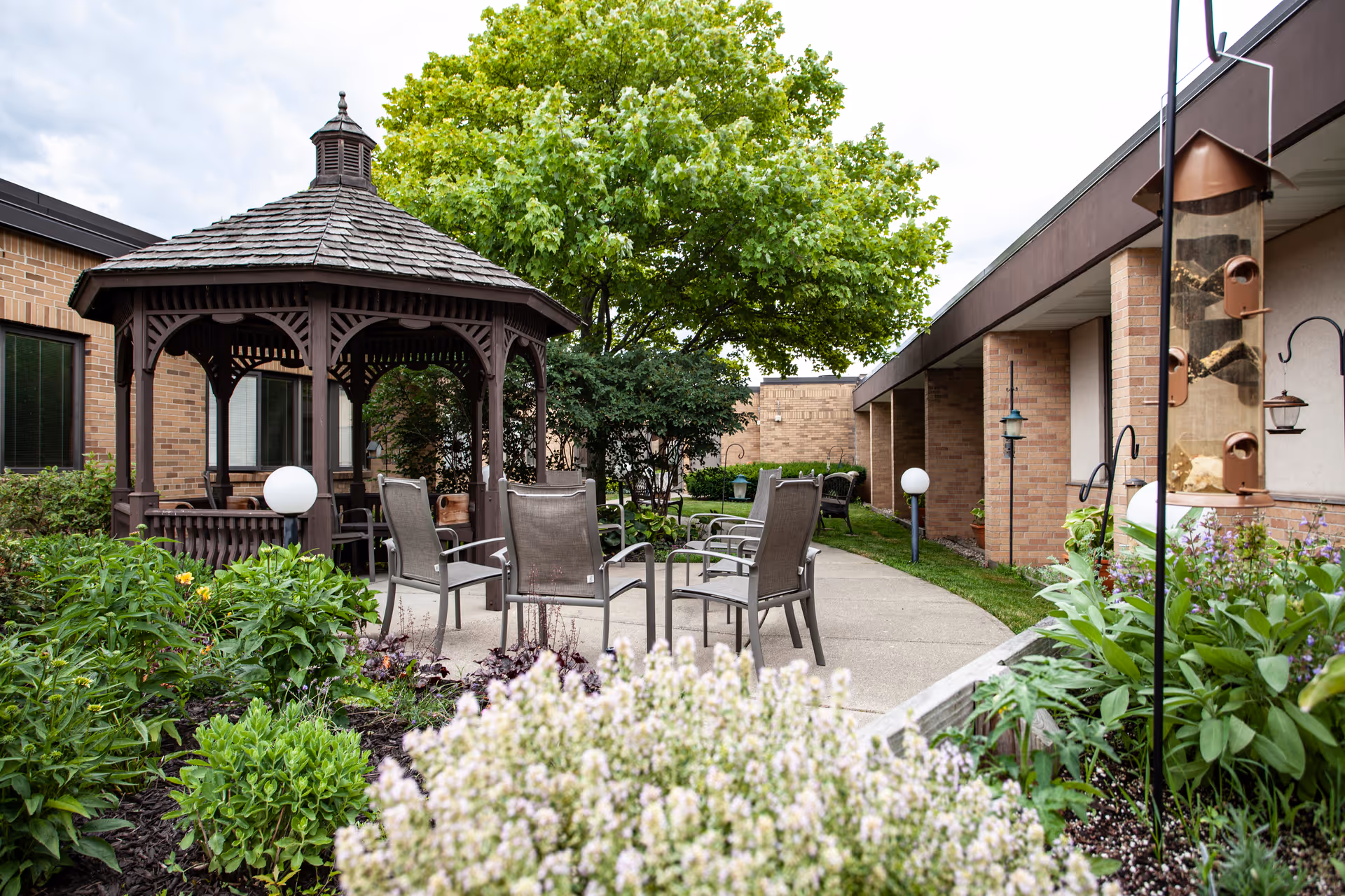Courtyard featuring a wooden gazebo, patio chairs around a paved path and landscaped flower beds in front of a brick senior living building.