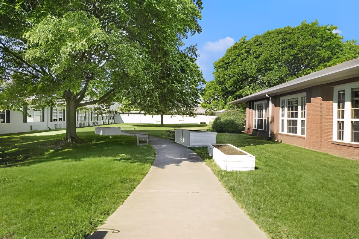 A paved walkway curves through a green lawn with large leafy trees providing shade. On the right side, there is a single-story brick building with multiple windows. Raised garden beds are placed along the path, and a bench is visible near the trees. The sky is clear and blue.