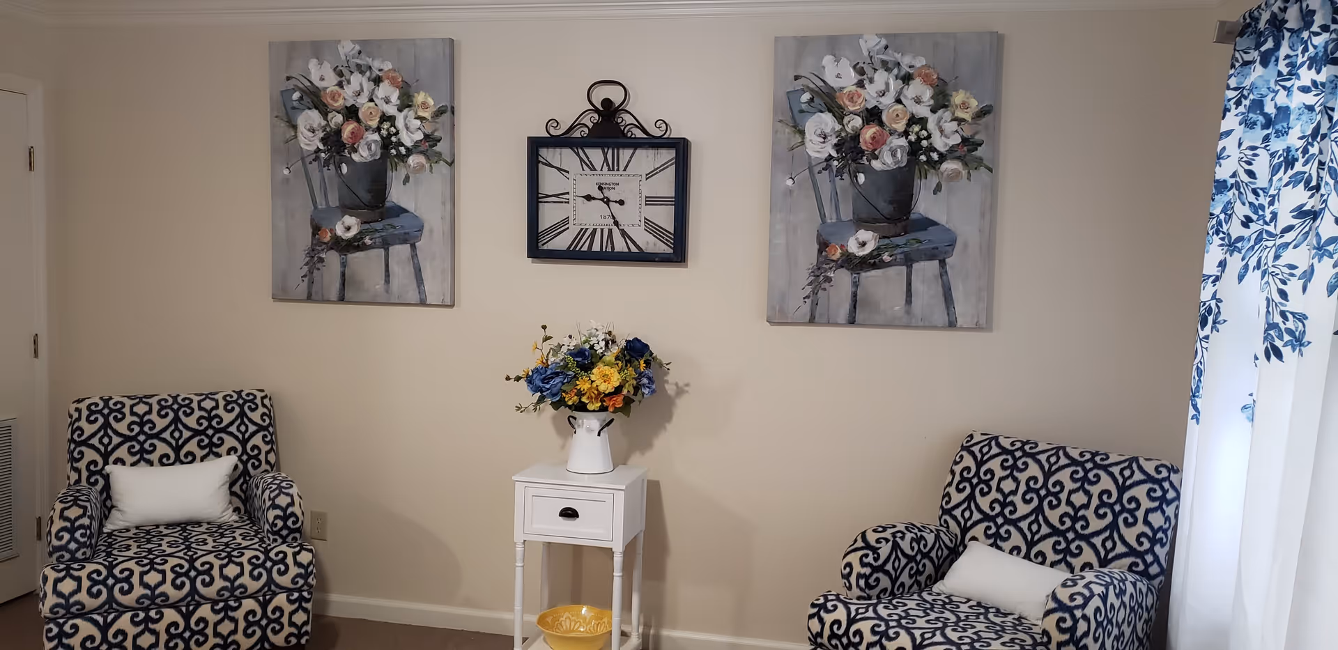 A cozy sitting area with two patterned armchairs featuring blue and white designs, each with a white lumbar pillow. Between the chairs is a small white side table holding a vase with a colorful bouquet of flowers. Above the table, a wall clock with Roman numerals is centered between two paintings of flower arrangements on chairs. To the right, a window with white curtains decorated with blue floral patterns lets in natural light.