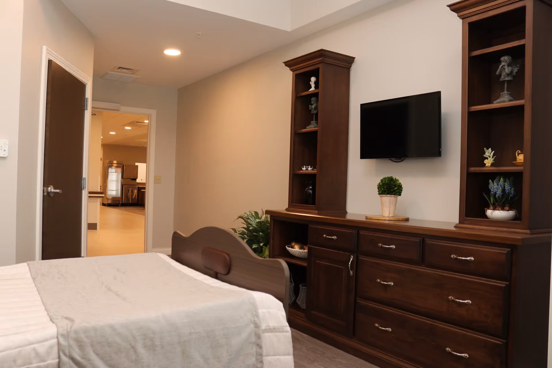 A furnished bedroom with a bed, large wooden dresser and shelving topped by a wall-mounted TV, and an open door leading to a hallway.