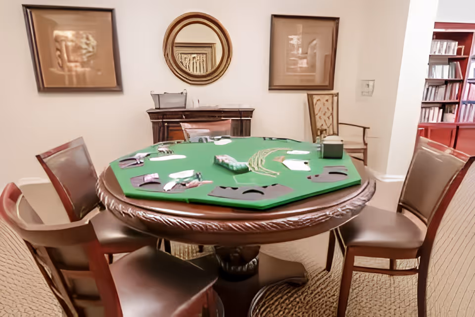 Round wooden card table with a green felt playing surface surrounded by chairs in a sitting room with framed art, a mirror, and bookshelves.