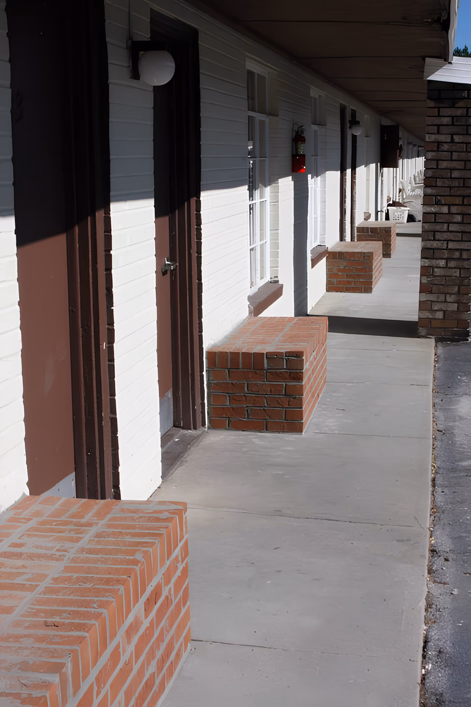 Outdoor corridor of a building with white walls and brown doors, featuring brick benches along the walkway and a fire extinguisher mounted on the wall.