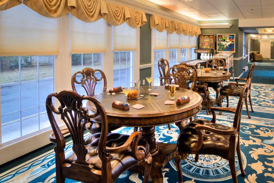 A well-lit room with large windows covered by beige blinds, featuring two round wooden tables with ornate carved chairs around them. The tables have drinks, snacks, and napkins placed on them. The room has a blue patterned carpet and pinball machines are visible in the background near a hallway.