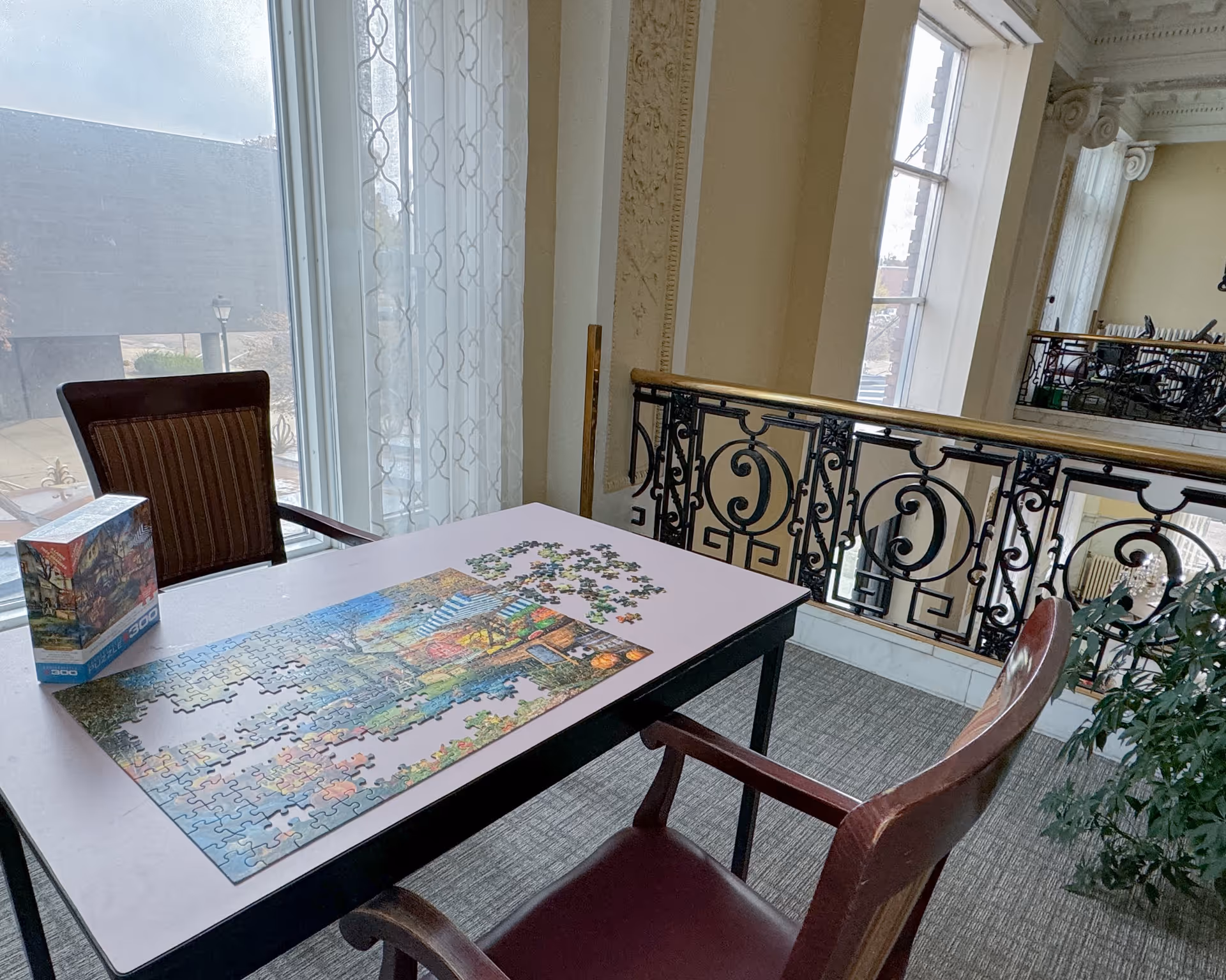 A table with a partially completed 300-piece jigsaw puzzle set up near a window with sheer curtains. Two wooden chairs with armrests are positioned on either side of the table. The room features ornate molding and a decorative black and gold railing overlooking a lower level.