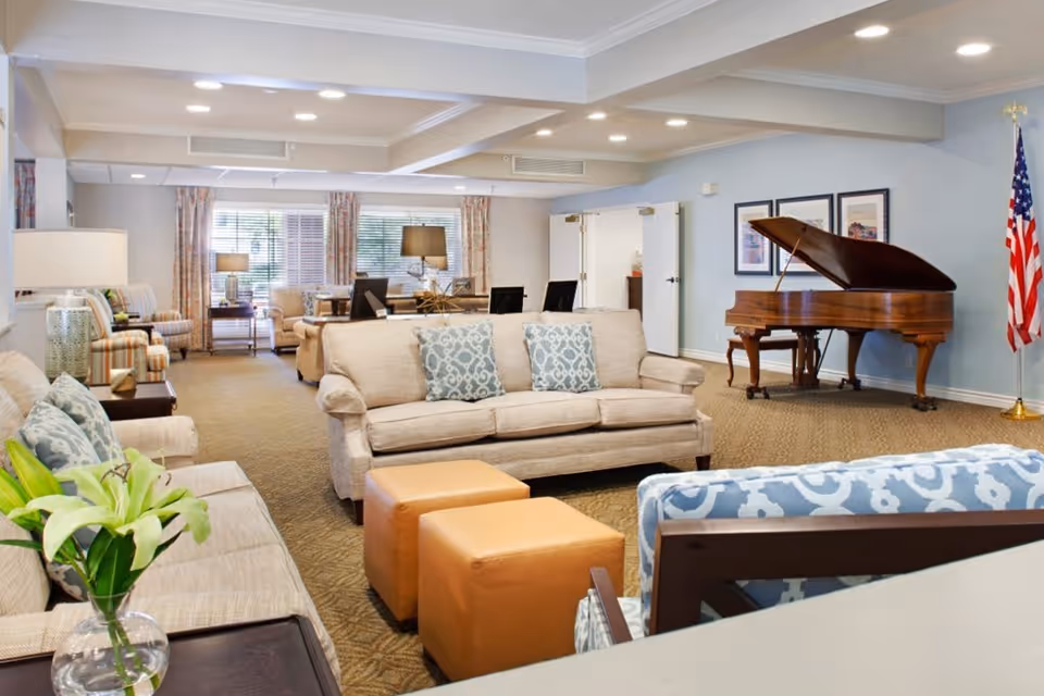A spacious and well-lit living room area in a senior living facility featuring beige sofas with patterned cushions, two tan ottomans, a blue patterned armchair, a grand piano near the wall with framed artwork above it, an American flag in the corner, and large windows with curtains allowing natural light to fill the room.