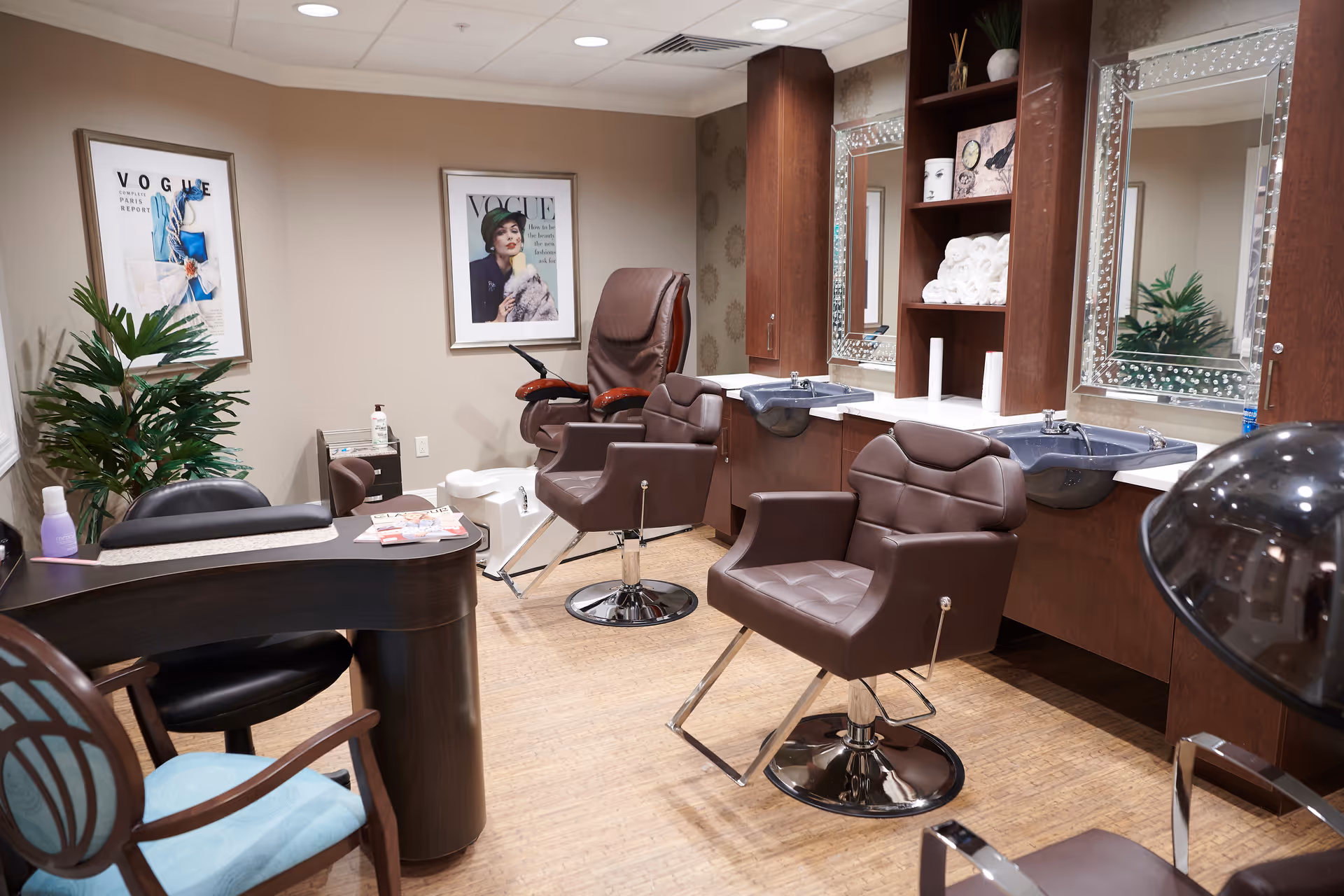 Interior of a salon area in a senior living facility featuring two brown salon chairs in front of sinks with large mirrors, a manicure table with chairs, framed Vogue magazine covers on the wall, and a potted plant in the corner.