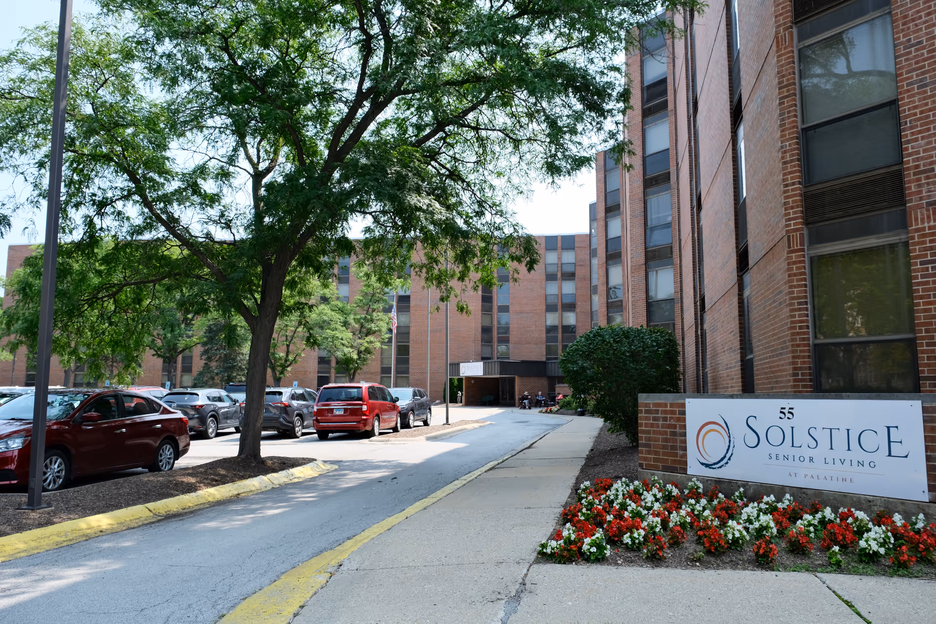 Driveway and entrance of a brick senior living building with parked cars, trees, and a 'Solstice Senior Living' sign by a flowerbed.