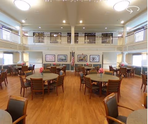 Spacious two-story dining room with round tables and chairs, hardwood floors, a second-floor balcony, and framed artwork on the far wall.