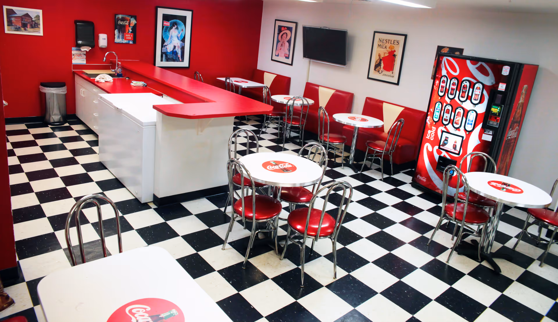 A retro-style dining area with black and white checkered floor tiles, red and white tables and chairs, a red countertop with a sink, and a Coca-Cola vending machine. The walls are decorated with vintage posters and a mounted TV.