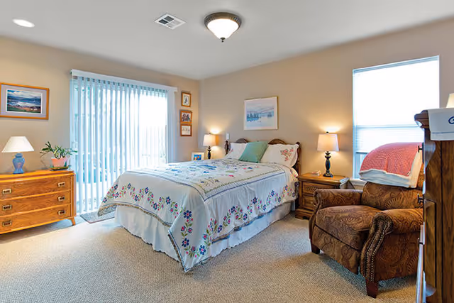 A cozy bedroom with a bed covered in a floral quilt, two bedside tables with lamps, a wooden dresser with a lamp and plant, an upholstered armchair with a folded blanket on it, and large windows with vertical blinds allowing natural light to enter.