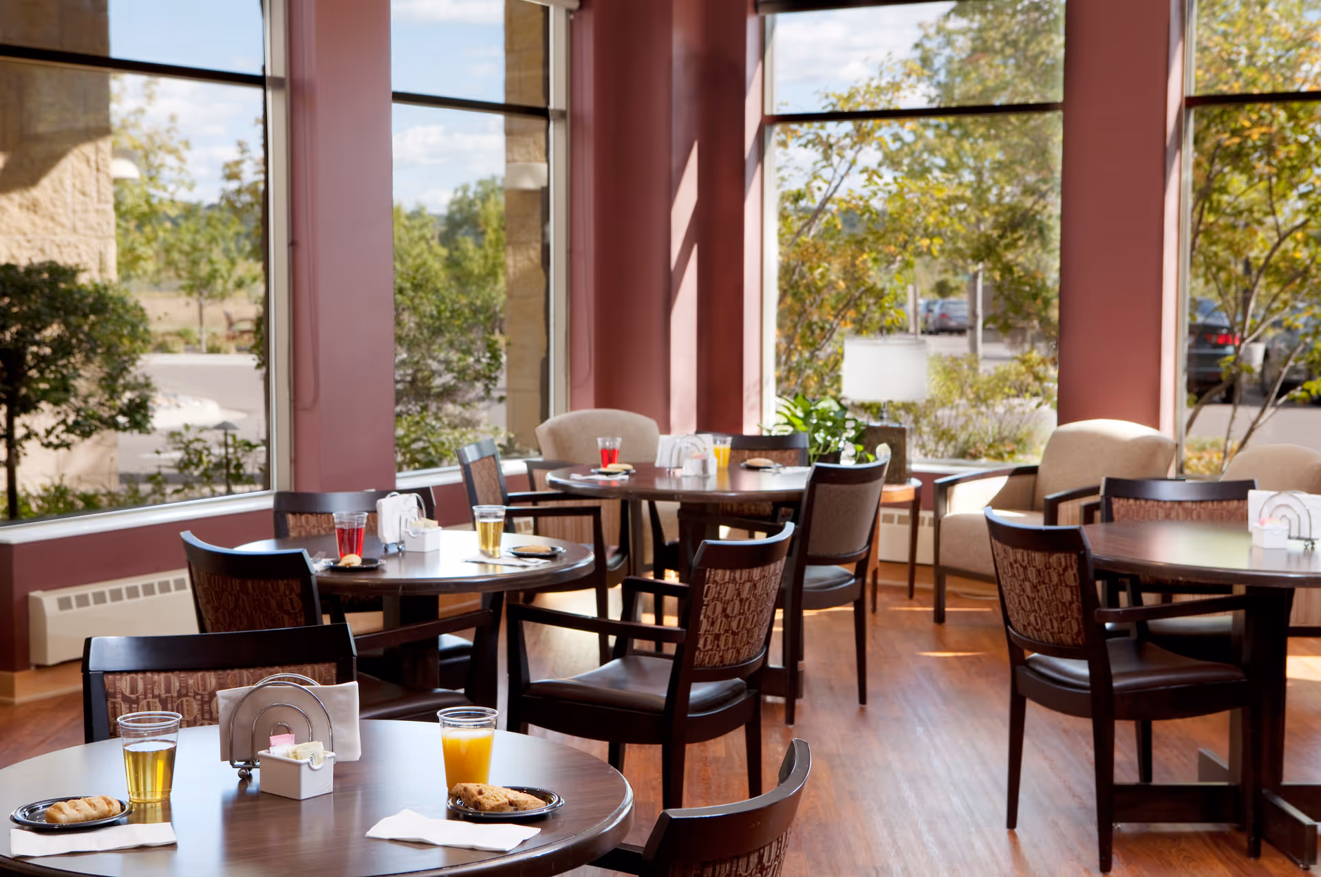 Dining room with round wooden tables set with drinks and pastries beside large windows overlooking trees.