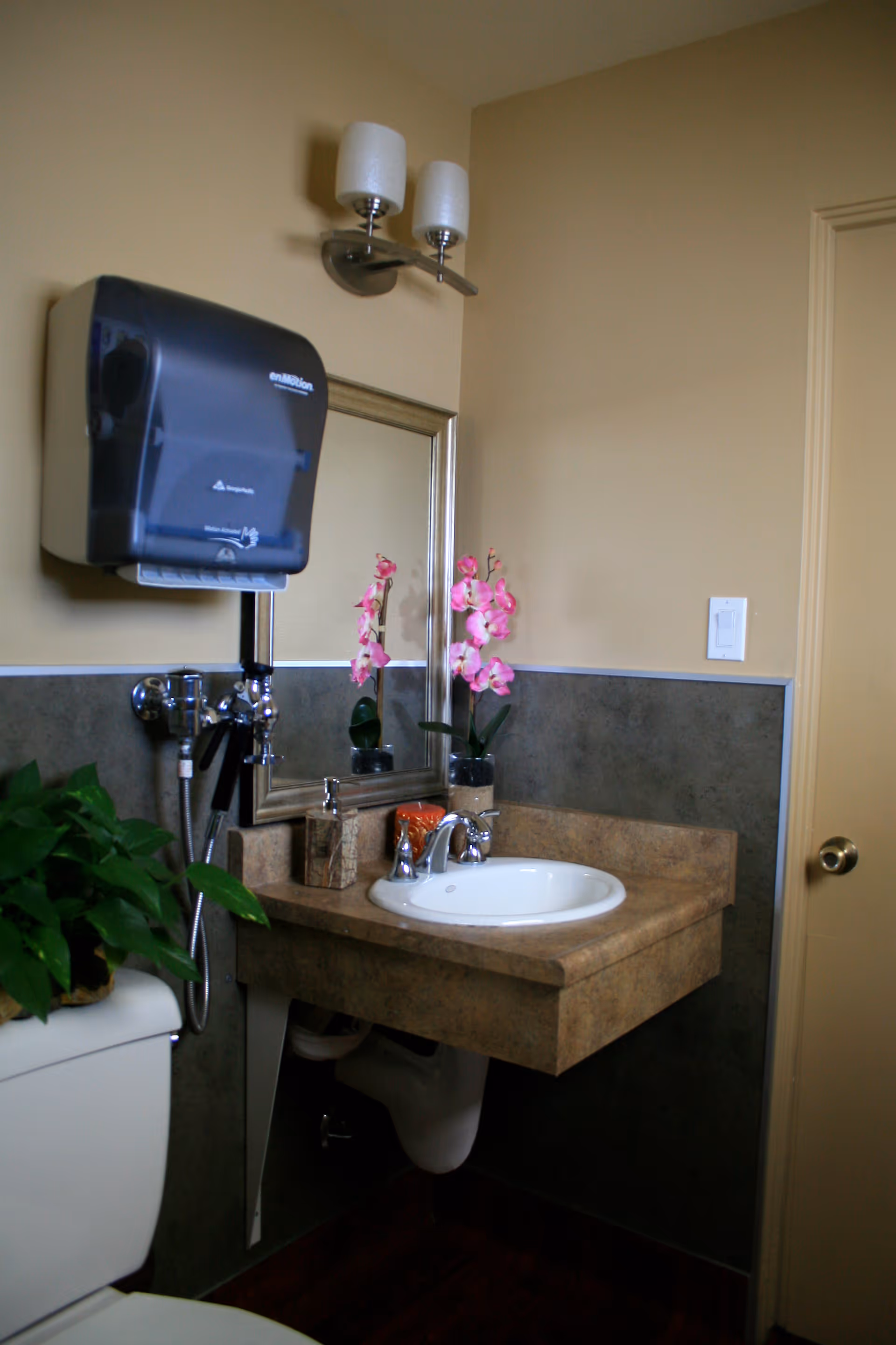 A small bathroom sink area with a beige countertop and a white sink. Above the sink is a rectangular mirror with a silver frame and a two-light wall fixture with frosted glass shades. To the left of the sink is a black paper towel dispenser mounted on the wall. There is a pink orchid plant in a pot on the countertop and a green leafy plant on top of a white toilet tank. The walls are painted beige with a darker wainscoting panel. A door with a brass knob is visible on the right side.