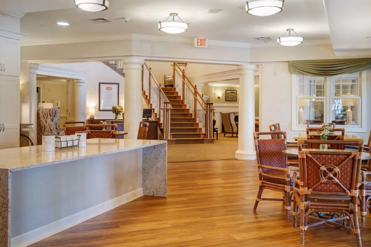 Bright communal dining and lounge area with a marble-topped island, wooden tables and chairs, and a central staircase.