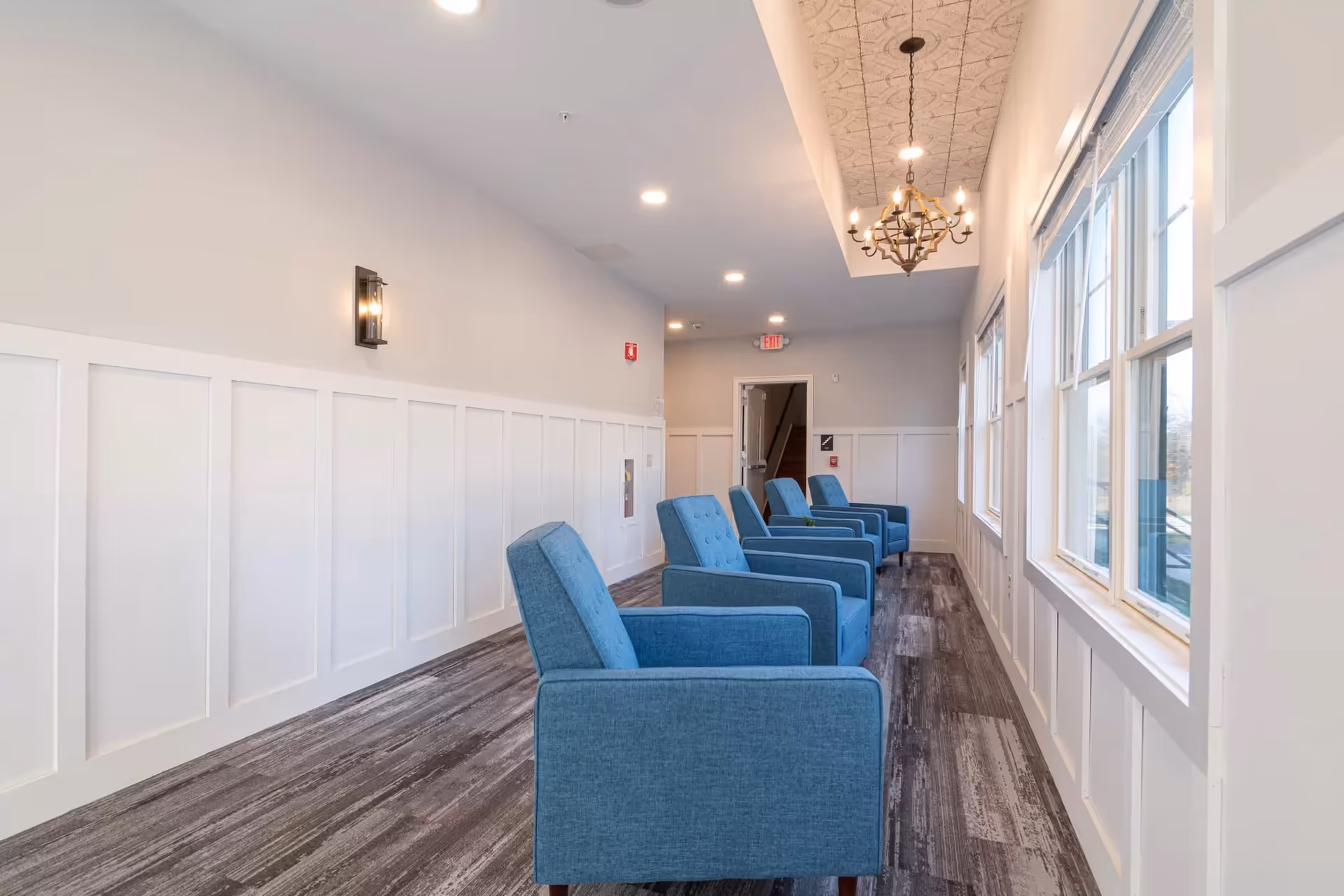 A bright hallway with large windows on one side and white paneled walls on the other. Four blue upholstered armchairs are lined up facing the windows. The ceiling features recessed lighting and a decorative chandelier. The floor has a wood-like textured finish.