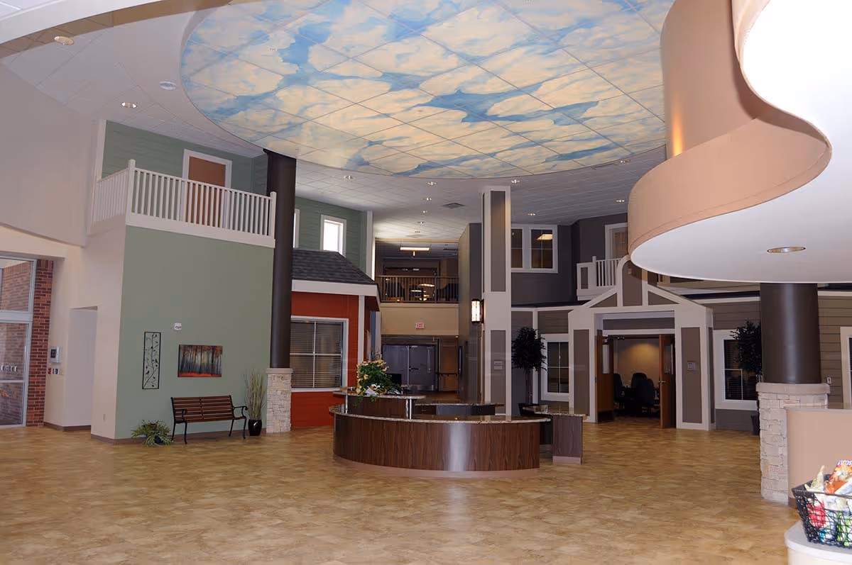 Spacious interior lobby area of a senior living facility with a circular wooden reception desk in the center, decorative small house-like structures along the walls, a bench with wall art on the left, and a ceiling painted with a sky and clouds design.