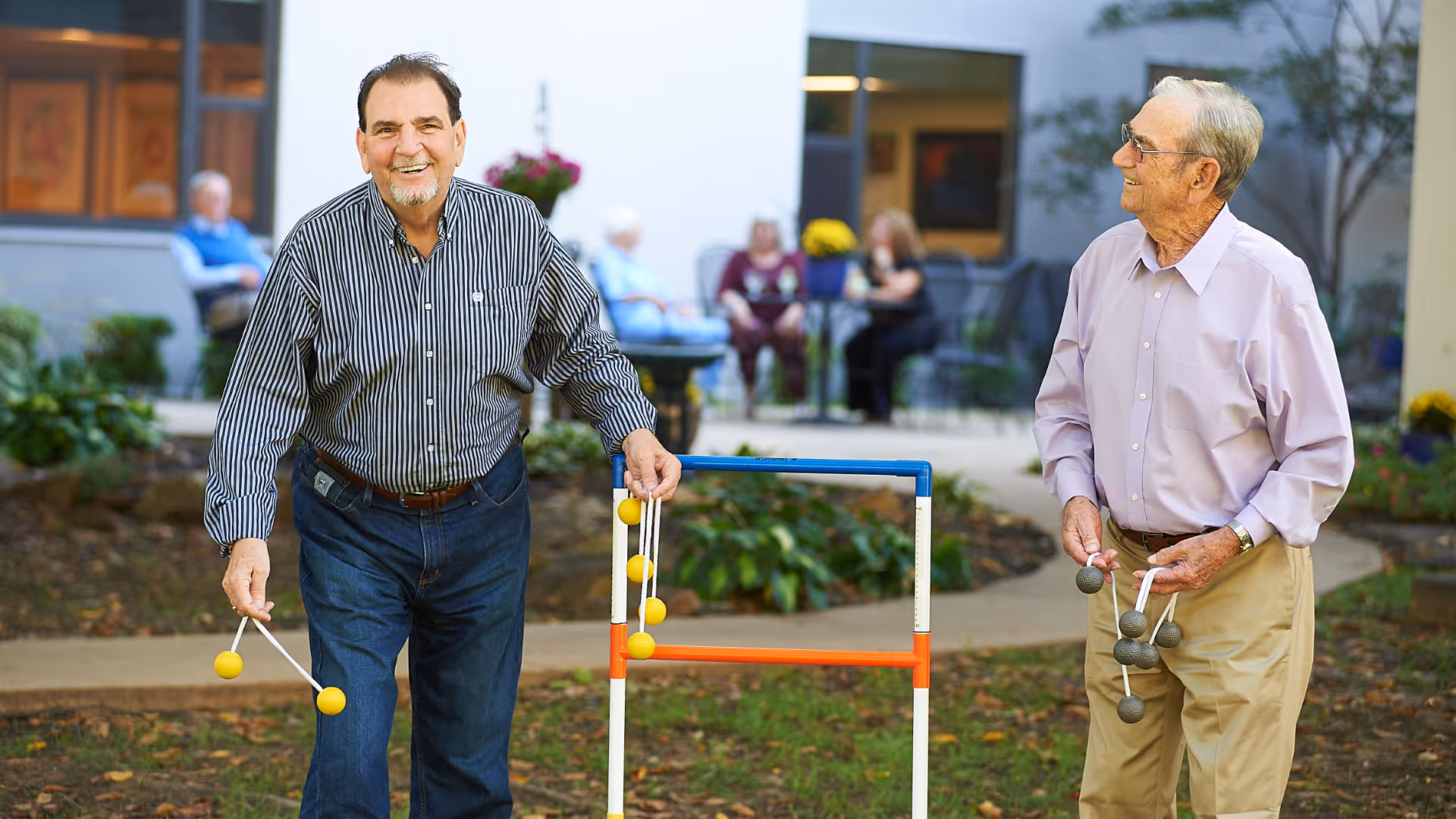 Two elderly men playing a game of ladder toss outdoors in a garden area of a senior living facility. One man is smiling and holding yellow bolas, while the other man is looking at him and holding gray bolas. In the background, other seniors are seated and socializing near the building.