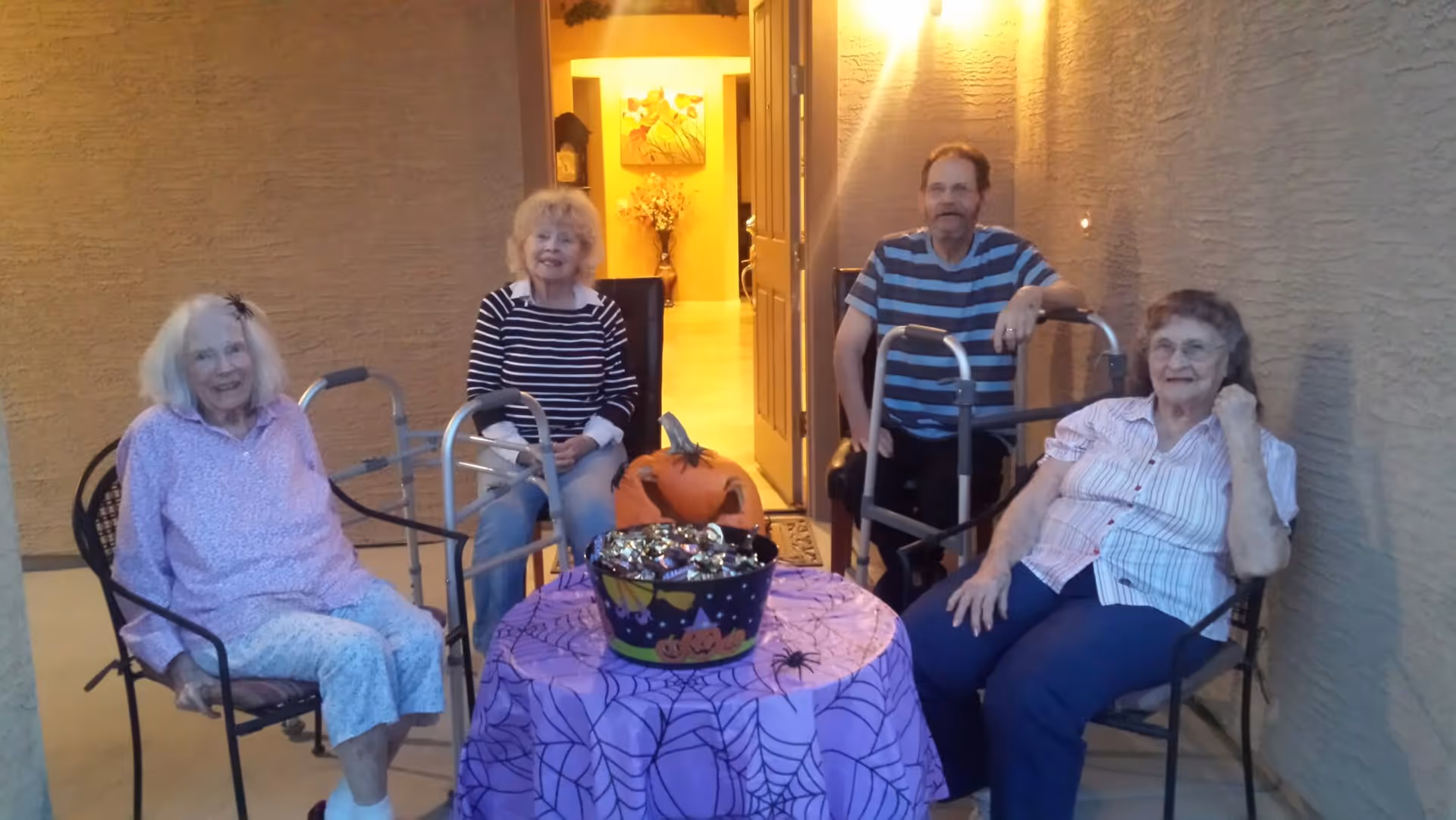 Four elderly individuals sitting outside on chairs around a small table covered with a purple Halloween-themed tablecloth featuring spiderwebs. On the table is a bucket filled with candy and a carved pumpkin. Two of the individuals have walkers. The setting appears to be a porch or patio area with a door open to a warmly lit interior.