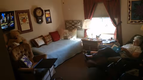 A cozy room with a single bed covered with a light blue blanket and several pillows. There is a brown armchair with a laptop on a tray in front of it. On the wall above the bed are framed pictures and a clock. A window with red curtains lets in natural light. A wooden side table with a lamp and various items sits next to the window. An elderly person is sitting in a recliner chair near the window, reading or looking at something in their hands.