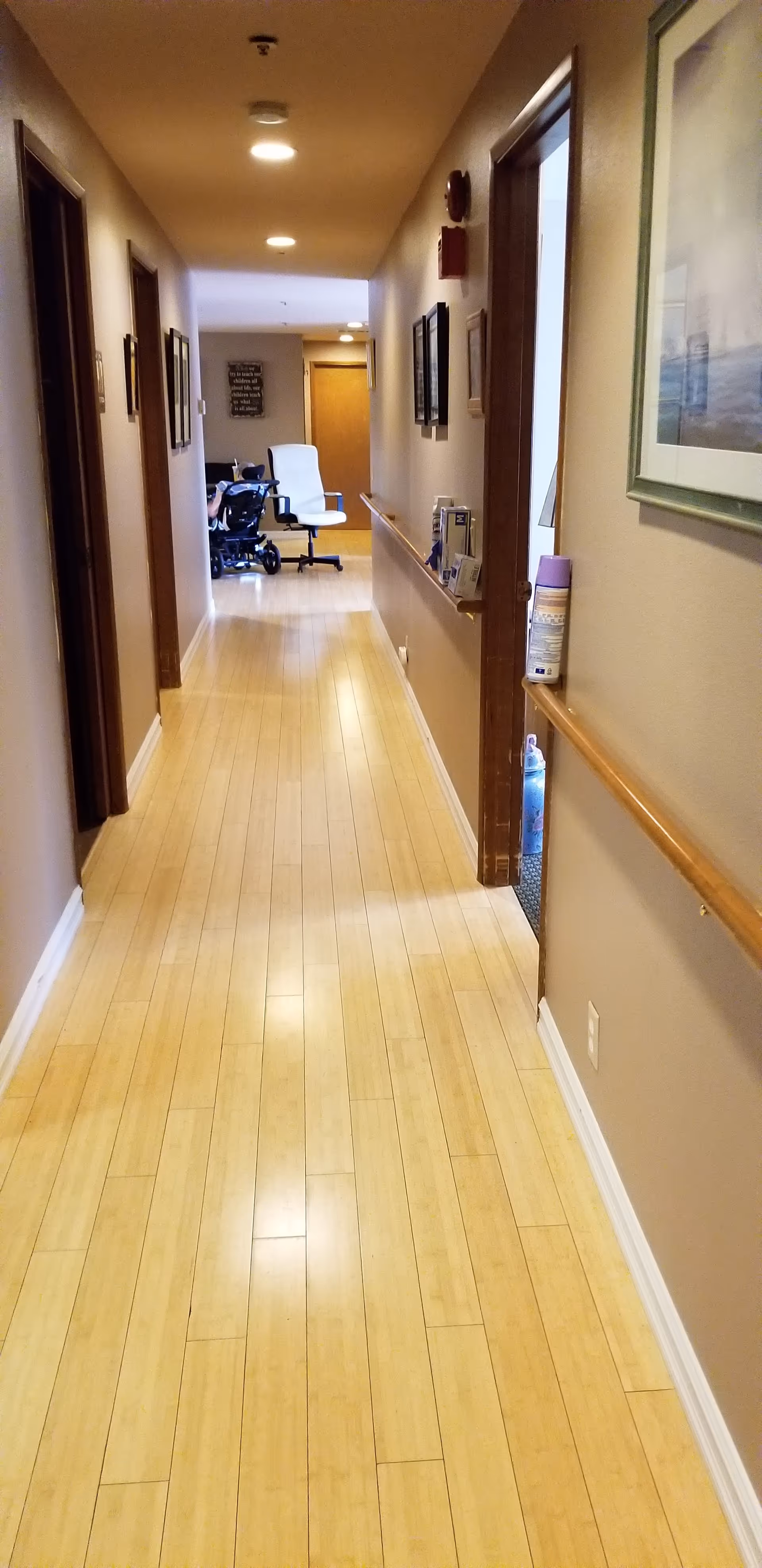 A long, narrow hallway with light wood flooring and beige walls. Several doorways and framed pictures line the walls. At the end of the hallway, there is a white office chair and a person in a wheelchair. The hallway is well-lit with ceiling lights.