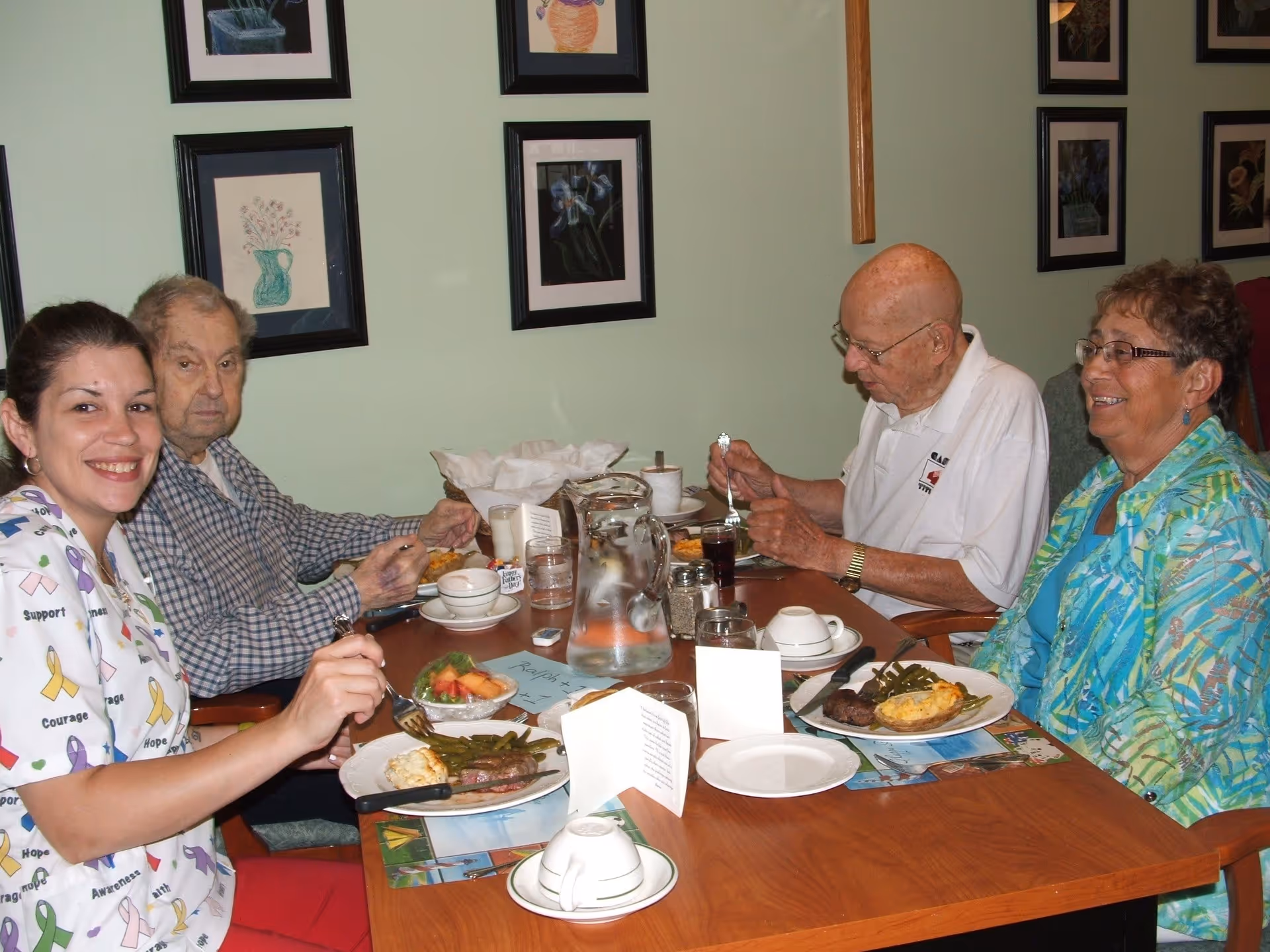 Four people sitting around a wooden dining table enjoying a meal together in a room with light green walls decorated with framed floral artwork. The group includes two elderly men, one elderly woman, and a younger woman wearing a colorful shirt with awareness ribbons. Plates of food, glasses, and a water pitcher are on the table.