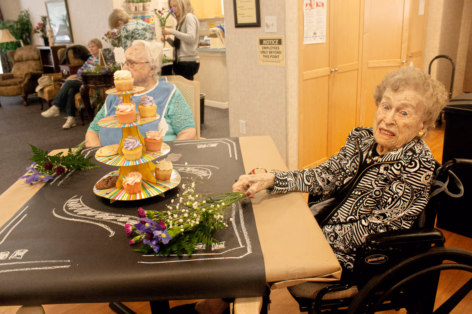 Two elderly women sitting at a table covered with black paper decorated with white chalk drawings. One woman in a wheelchair is holding a bouquet of flowers, and the other woman is seated nearby. A three-tiered stand with colorful cupcakes is on the table. In the background, other elderly people and a caregiver are visible in a cozy room with wooden cabinets and comfortable chairs.