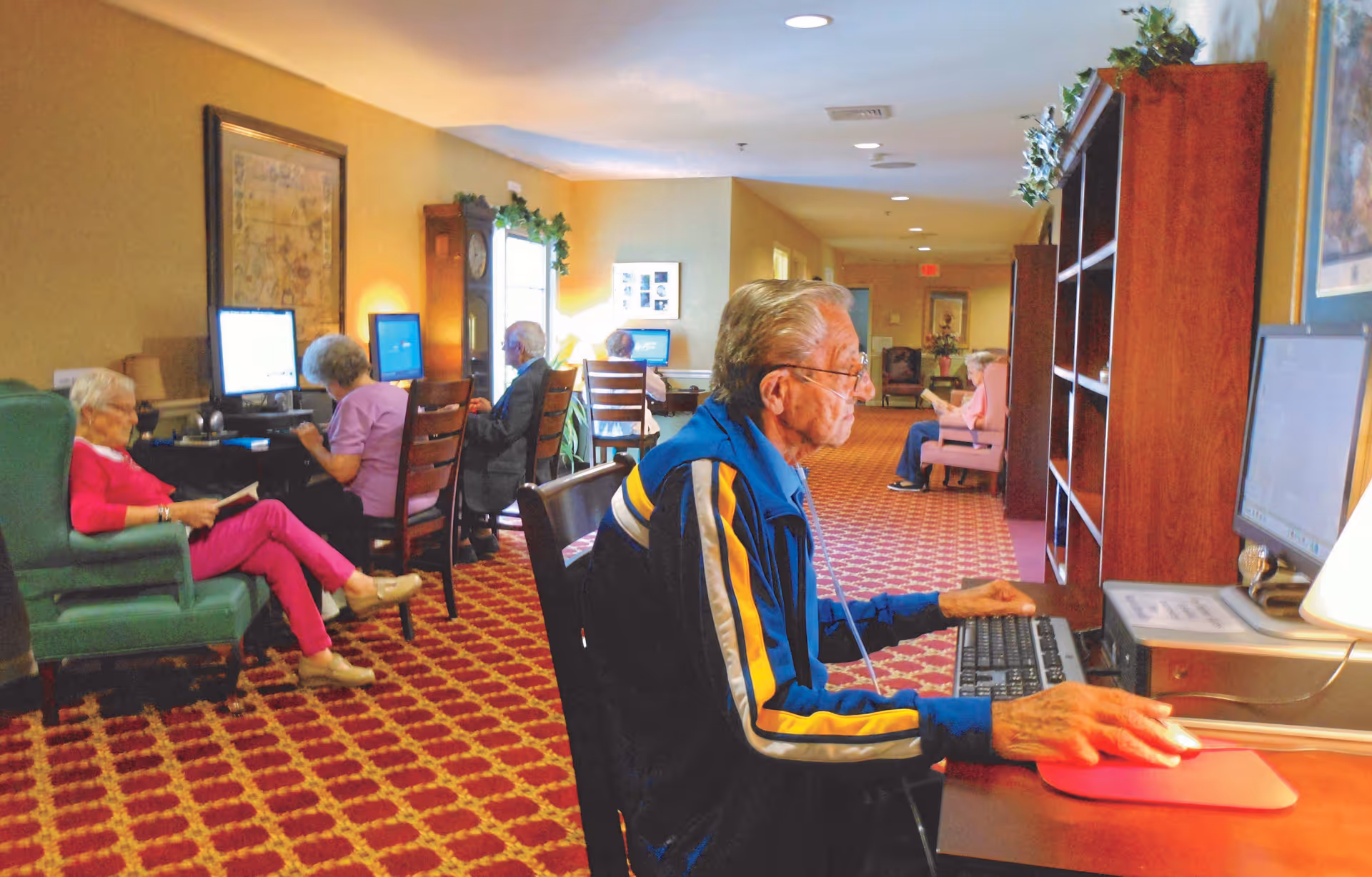 Several elderly individuals seated in a senior community computer room. One man in a blue and yellow jacket is using a desktop computer in the foreground, while others are seated at computers or reading in the background. The room has patterned red carpet, warm yellow walls, wooden furniture, and decorative plants.