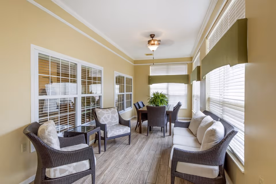 A bright sunroom with beige walls and large windows covered with white blinds. The room is furnished with a cushioned wicker sofa, two cushioned wicker armchairs, a small glass-top side table, and a wooden dining table with four wicker chairs. A ceiling fan with a light fixture hangs from the ceiling, and a green potted plant is placed on the dining table.