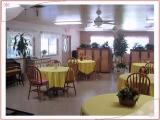 Interior view of a dining room in an assisted living facility with round tables covered in yellow tablecloths, wooden chairs, potted plants on tables and around the room, large windows with white curtains, and ceiling fans.