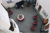 View from above of a nursing home common area with green carpet, four maroon armchairs arranged around a round table with a pink tablecloth and a flower arrangement. There is a piano against one wall, framed pictures on the walls, and a door leading to another room.