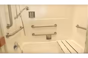 Accessible bathroom with a white bathtub equipped with multiple stainless steel grab bars and a foldable shower seat attached to the tub wall.