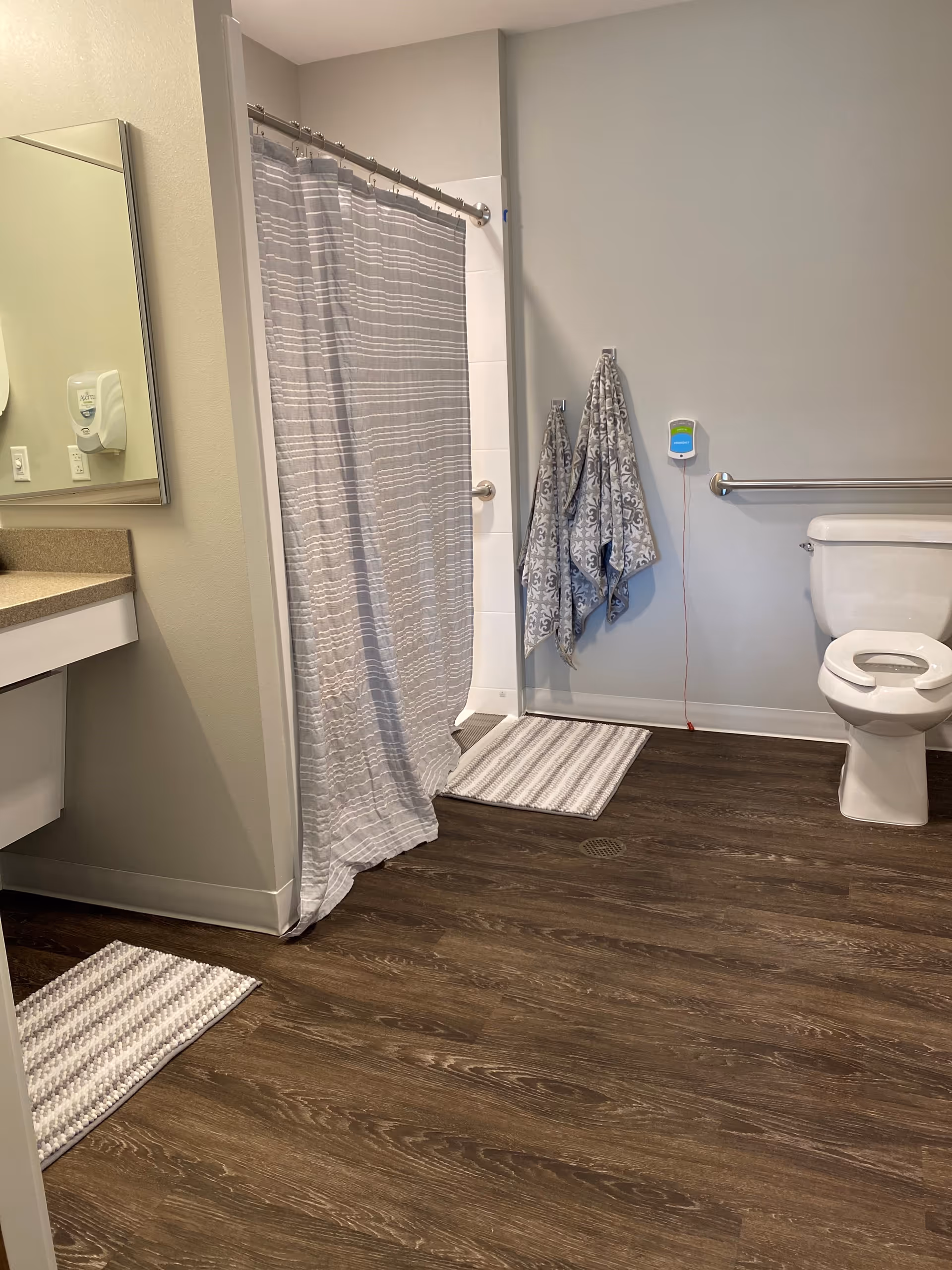 A bathroom with a toilet, a shower with a gray and white striped curtain, two patterned towels hanging on the wall, a sink with a mirror above it, and two striped floor mats on a wood-patterned floor.