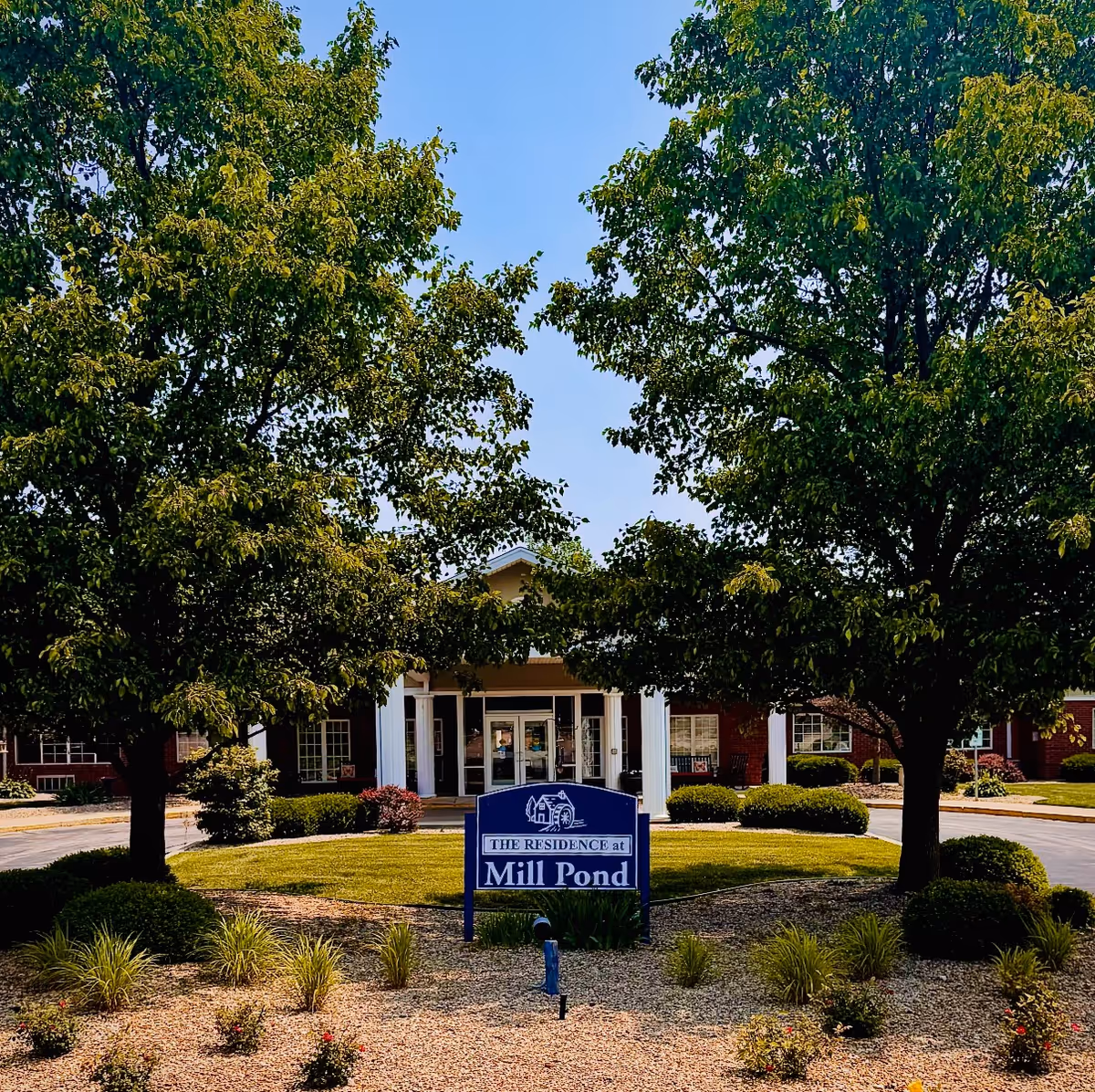 Front entrance of the Mill Pond building framed by trees, a landscaped lawn, and a sign reading 'Mill Pond'.