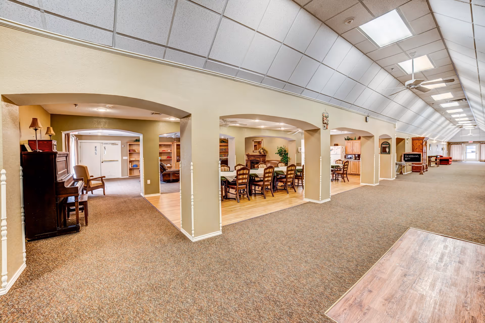 Interior view of a senior living facility hallway with carpeted floors and arched openings leading to various rooms including a dining area with tables and chairs, a sitting area with a piano and armchair, and a kitchen area. The ceiling has recessed lighting and ceiling fans, and there is a chalkboard sign indicating the direction to the restroom.