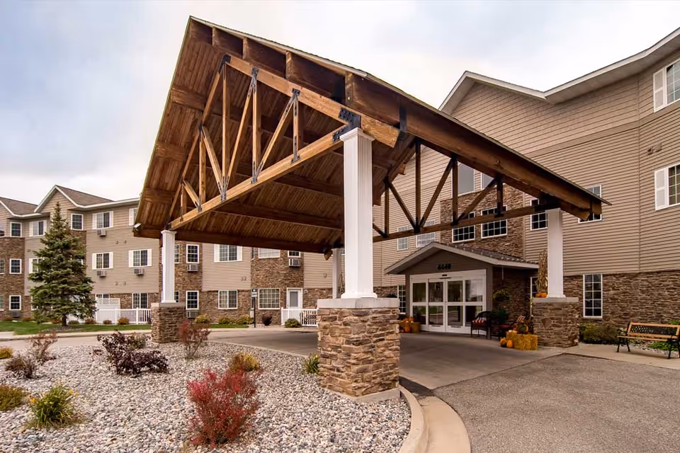 Entrance of a senior living facility named Edgewood Fargo featuring a large wooden covered driveway supported by stone and white pillars, with a beige multi-story building in the background. There are some small bushes and landscaping with rocks in the foreground.