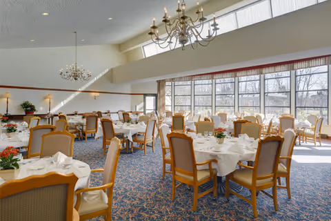 A bright dining room with multiple round tables covered with white tablecloths, set with plates, glasses, and napkins. The room features large windows letting in natural light, chandeliers hanging from the ceiling, and upholstered wooden chairs around each table. There are small flower arrangements on the tables and wall sconces providing additional lighting.