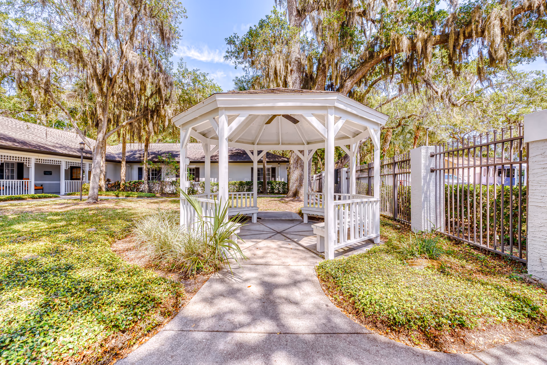 A white wooden gazebo with benches inside situated on a concrete path surrounded by green grass and plants. Large trees with hanging moss provide shade, and a building with a porch and white railing is visible in the background. A metal fence runs along the right side of the image under a clear blue sky.