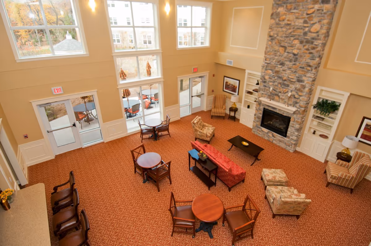 Sunlit two-story senior living common room with seating areas, round tables, and a stone fireplace.
