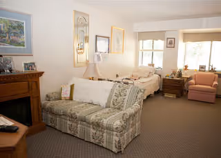 A cozy living room area with a floral patterned sofa, a wooden fireplace mantel with framed photos, a bed near the window, a wooden nightstand with a lamp, and a pink armchair. The room is softly lit with natural light coming through the windows.