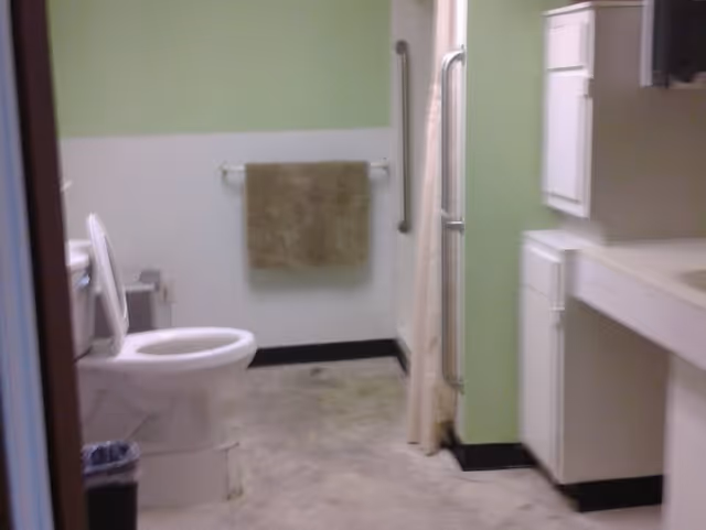 Bathroom with a white toilet, a beige towel hanging on a wall-mounted towel rack, a grab bar near the shower area with a curtain, and white cabinets with a countertop and sink. The walls are painted green and white, and the floor appears to be concrete or similar material.