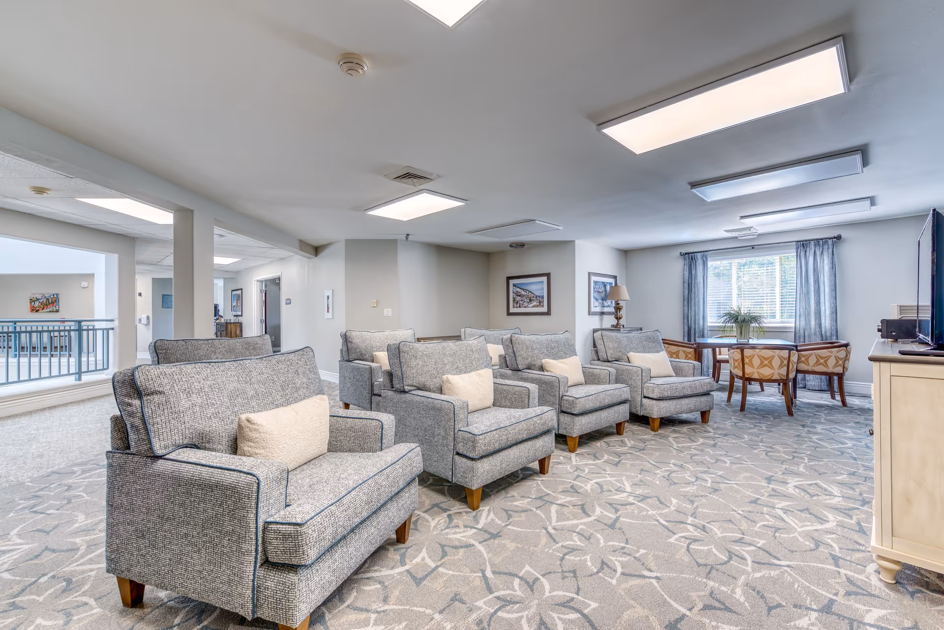 A bright and spacious senior living common area with four gray upholstered armchairs arranged in two rows facing a television on a white stand. The room has a patterned carpet, light gray walls, and a window with blue curtains letting in natural light. In the background, there is a small table with two chairs and framed artwork on the walls.