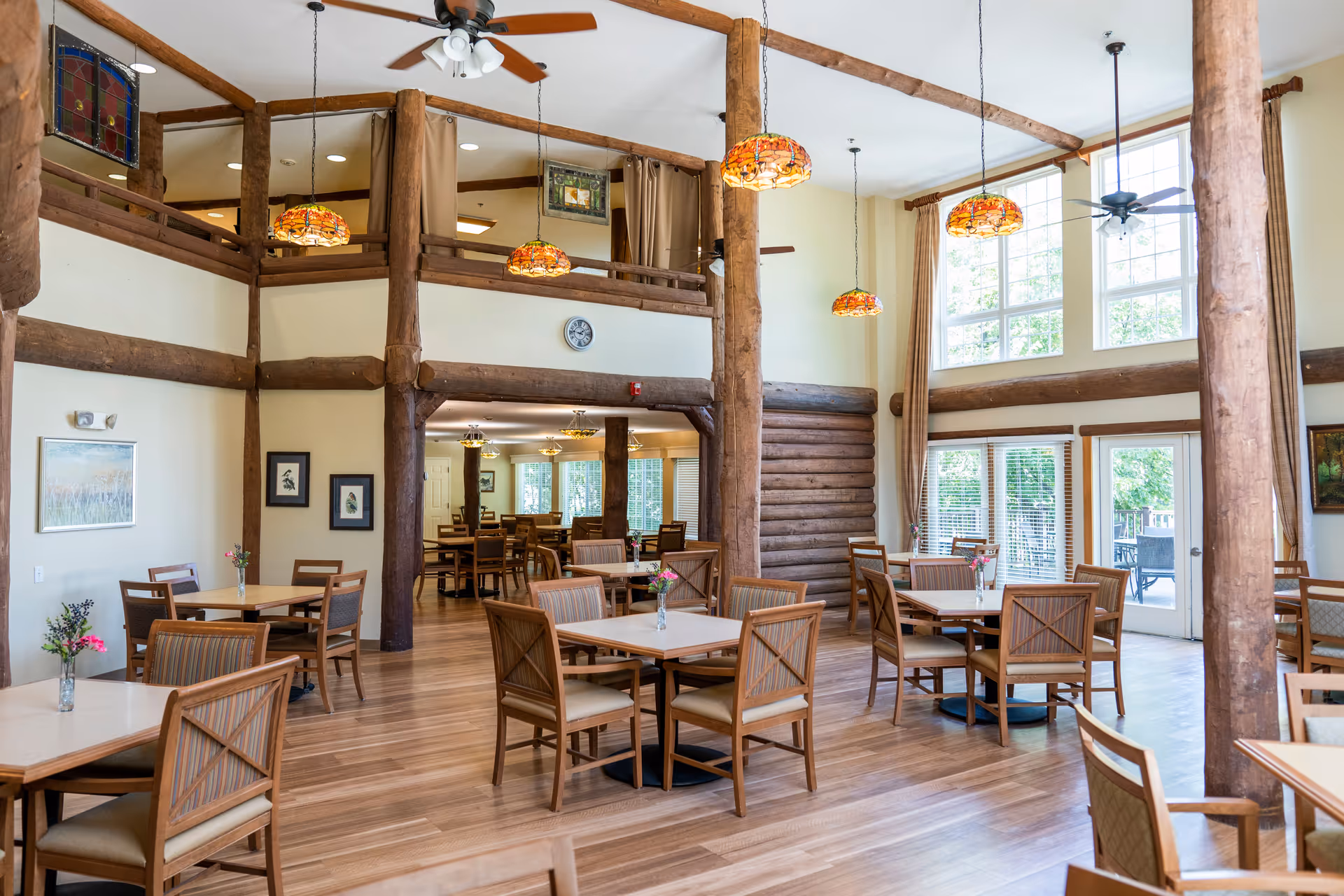 A spacious dining room with wooden floors and rustic wooden beams. Several square tables with chairs are arranged throughout the room, each table decorated with a small vase of flowers. Large windows allow natural light to fill the space, and stained glass pendant lights hang from the ceiling. There is a door leading to an outdoor patio area.