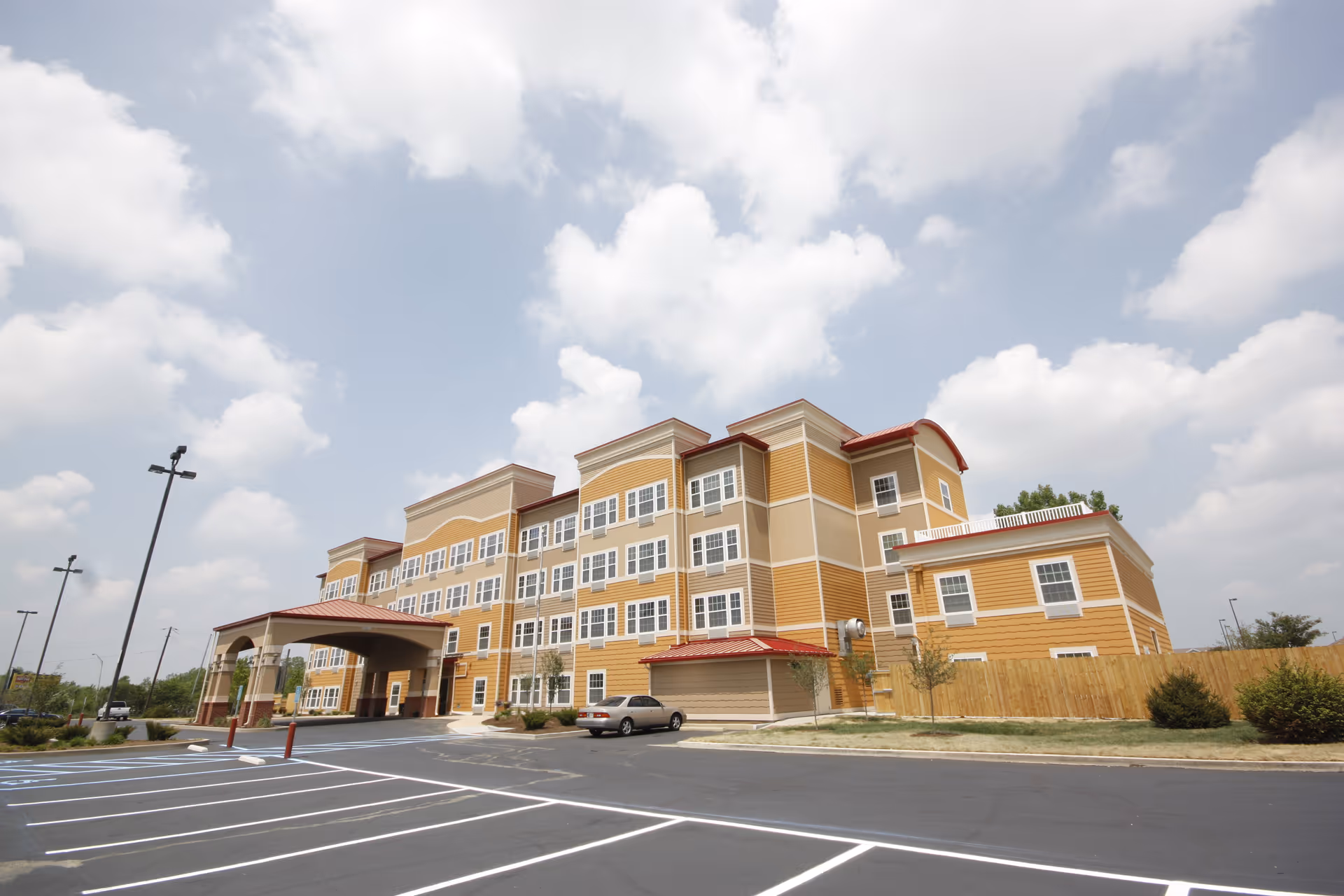 Exterior view of a multi-story senior living facility building with a covered entrance, multiple windows, a parking lot with marked spaces, and a partly cloudy sky.
