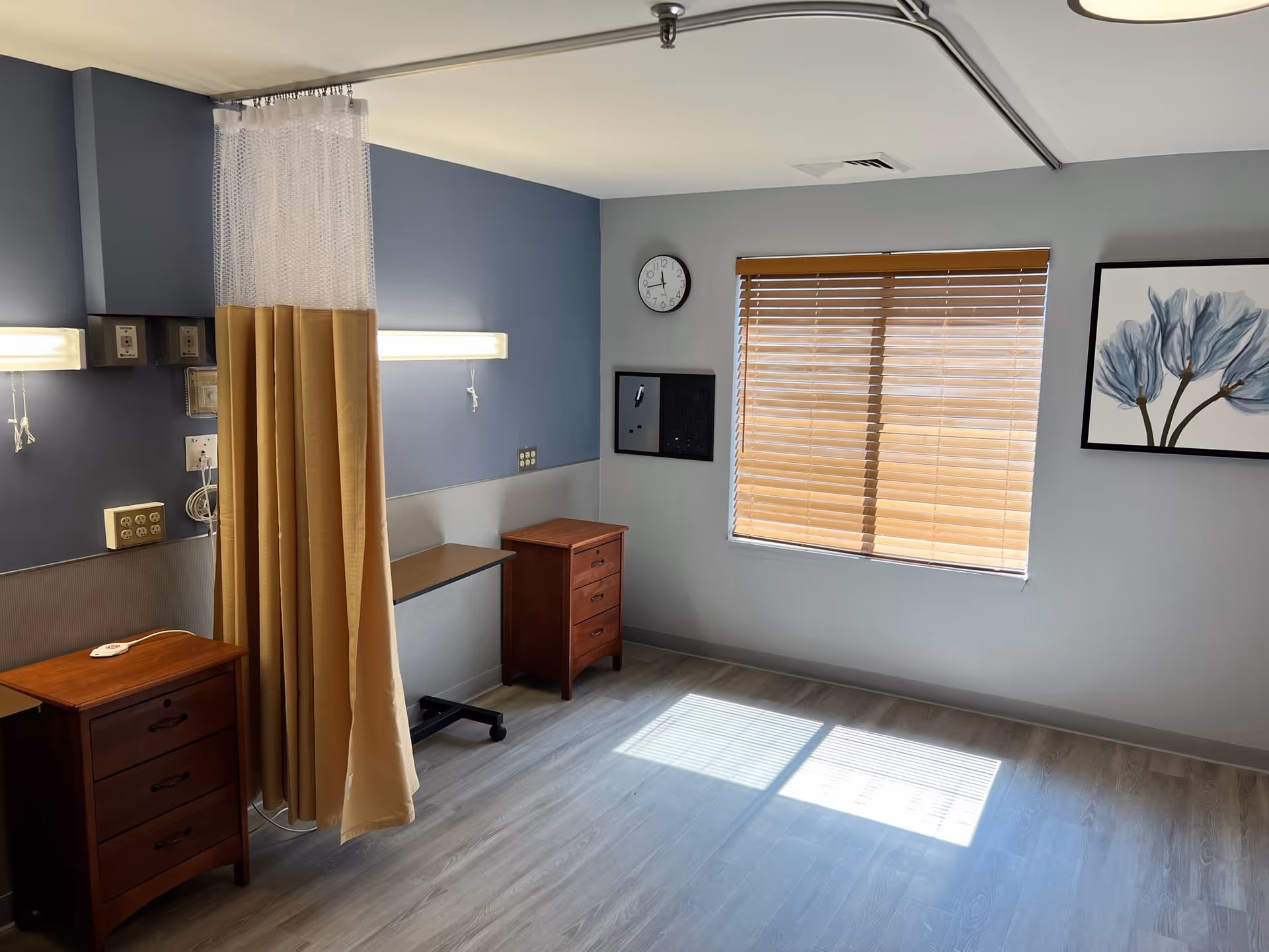 Empty nursing facility patient room with a privacy curtain, two wooden bedside cabinets, wall-mounted lights, a clock, and a window with blinds and floral artwork.