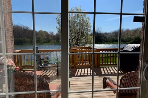 View through a glass-paned sliding door onto a wooden deck with patio chairs and a grill overlooking a lake and trees.